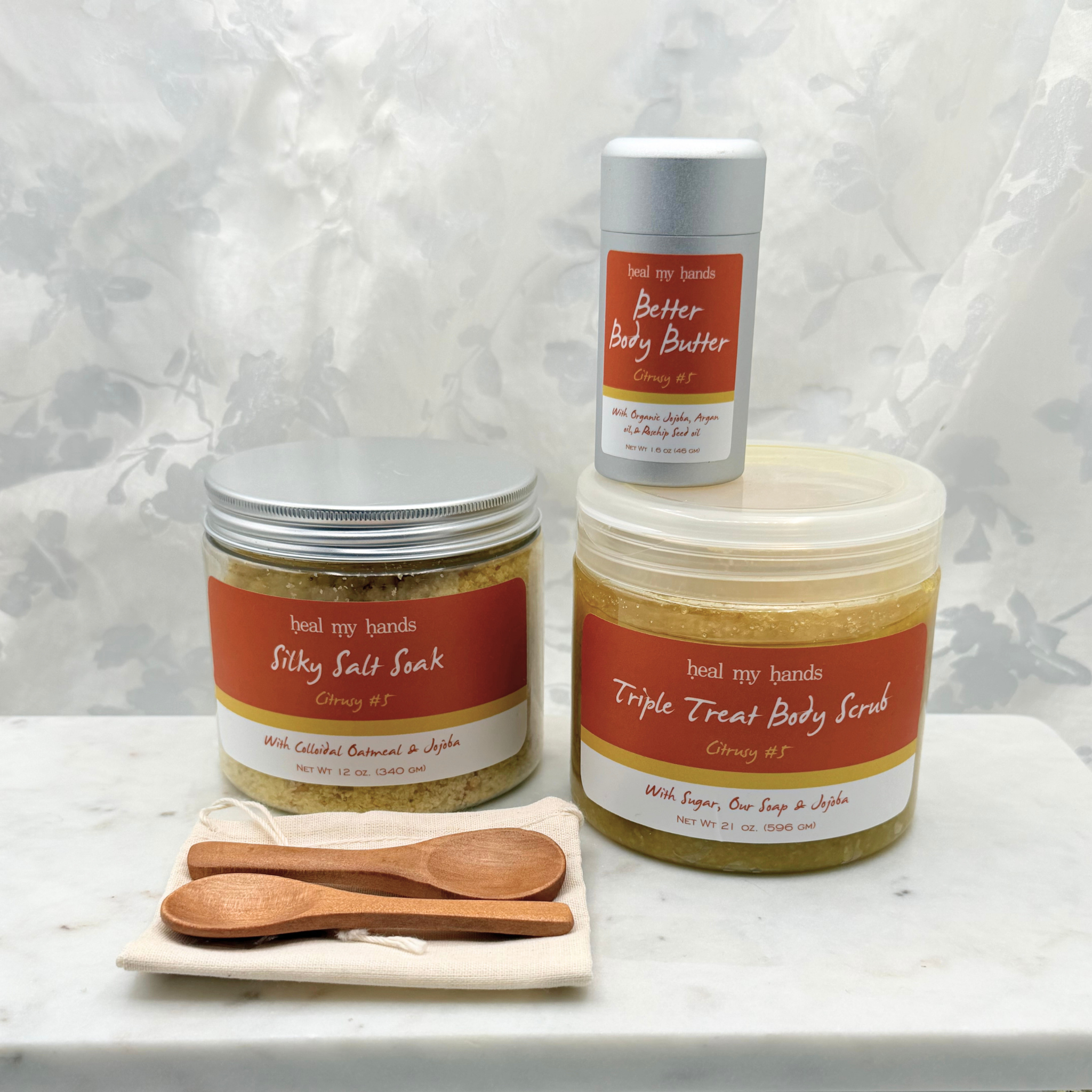 A jar of Silky Salt Soak, Triple Treat Body Scrub, and Better Body Butter in Citrusy #5 with two small wooden spoons and a muslin pouch on a marble surface.