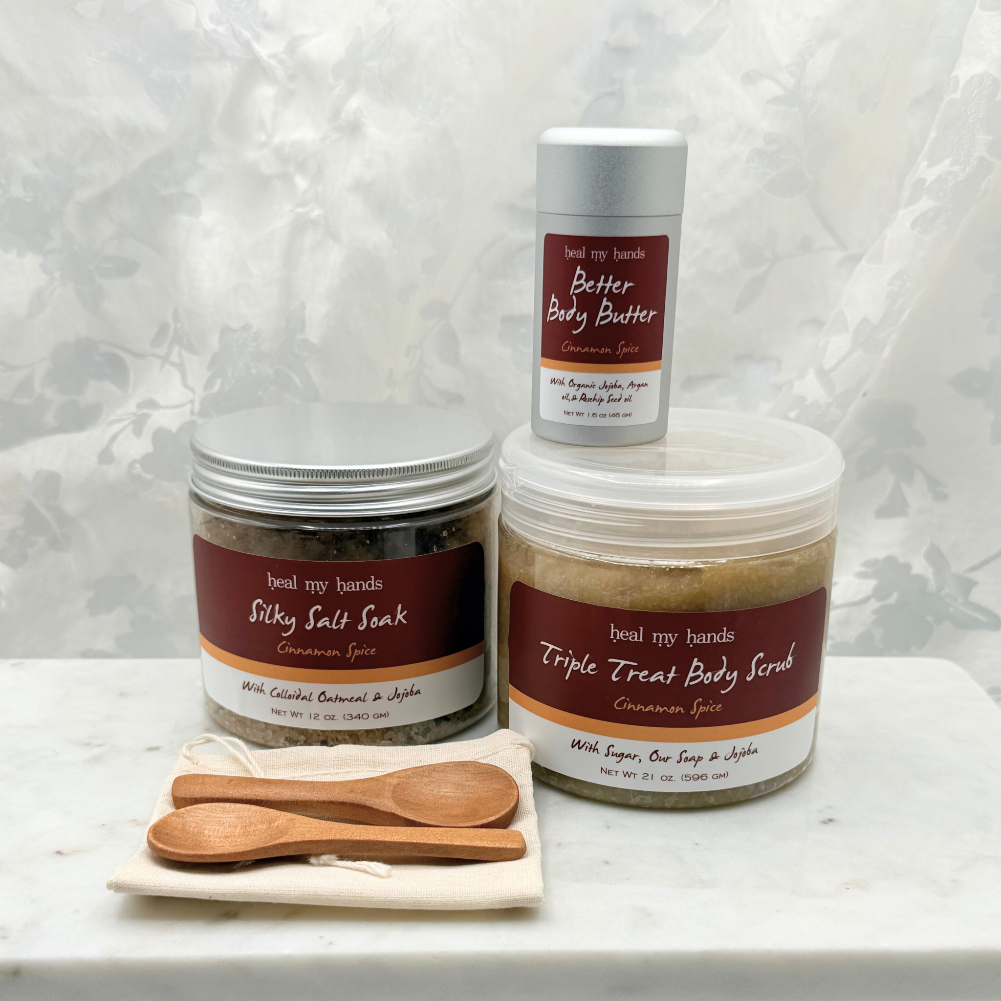 A jar of Silky Salt Soak, Triple Treat Body Scrub, and Better Body Butter in Cinnamon Spice with two small wooden spoons and a muslin pouch on a marble surface.