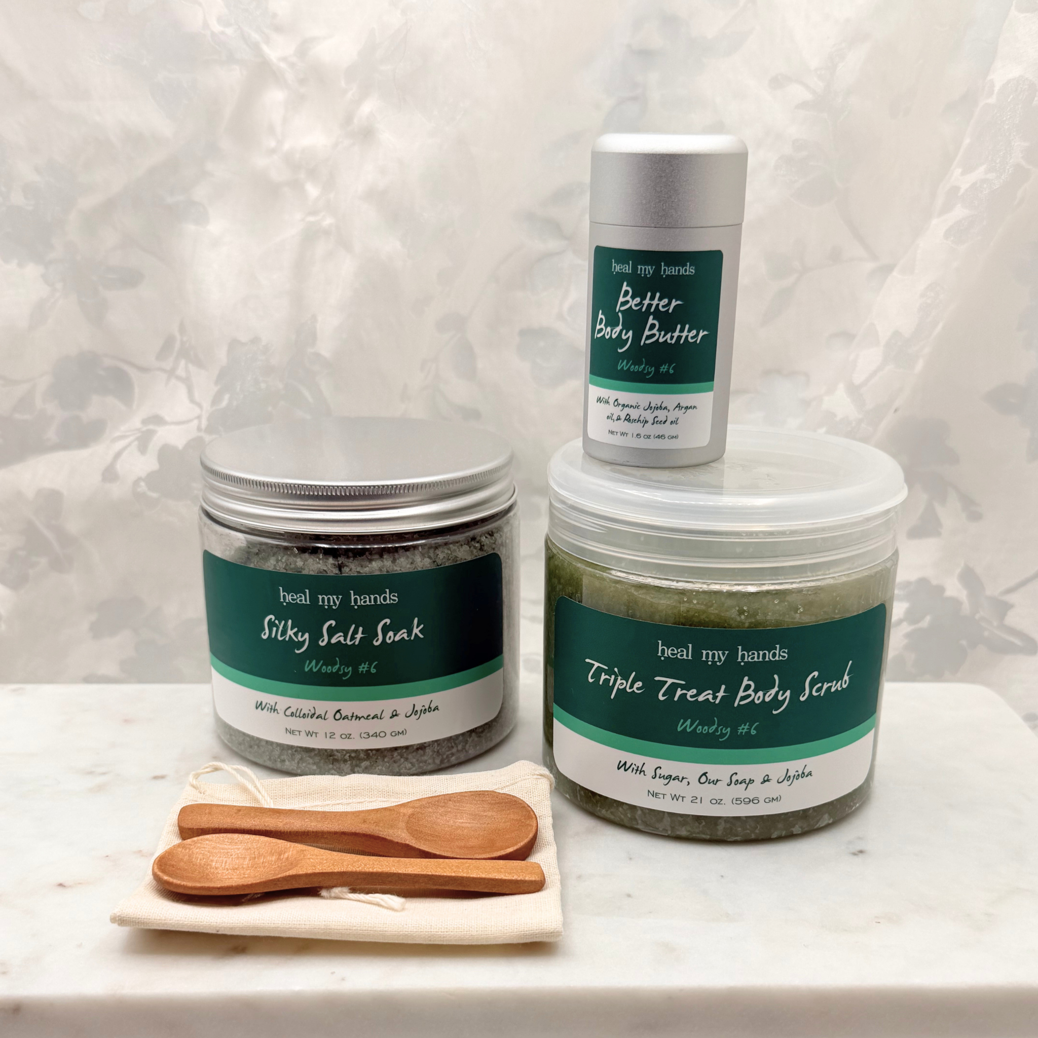 A jar of Silky Salt Soak, Triple Treat Body Scrub, and Better Body Butter in Woodsy #6 with two small wooden spoons and a muslin pouch on a marble surface.