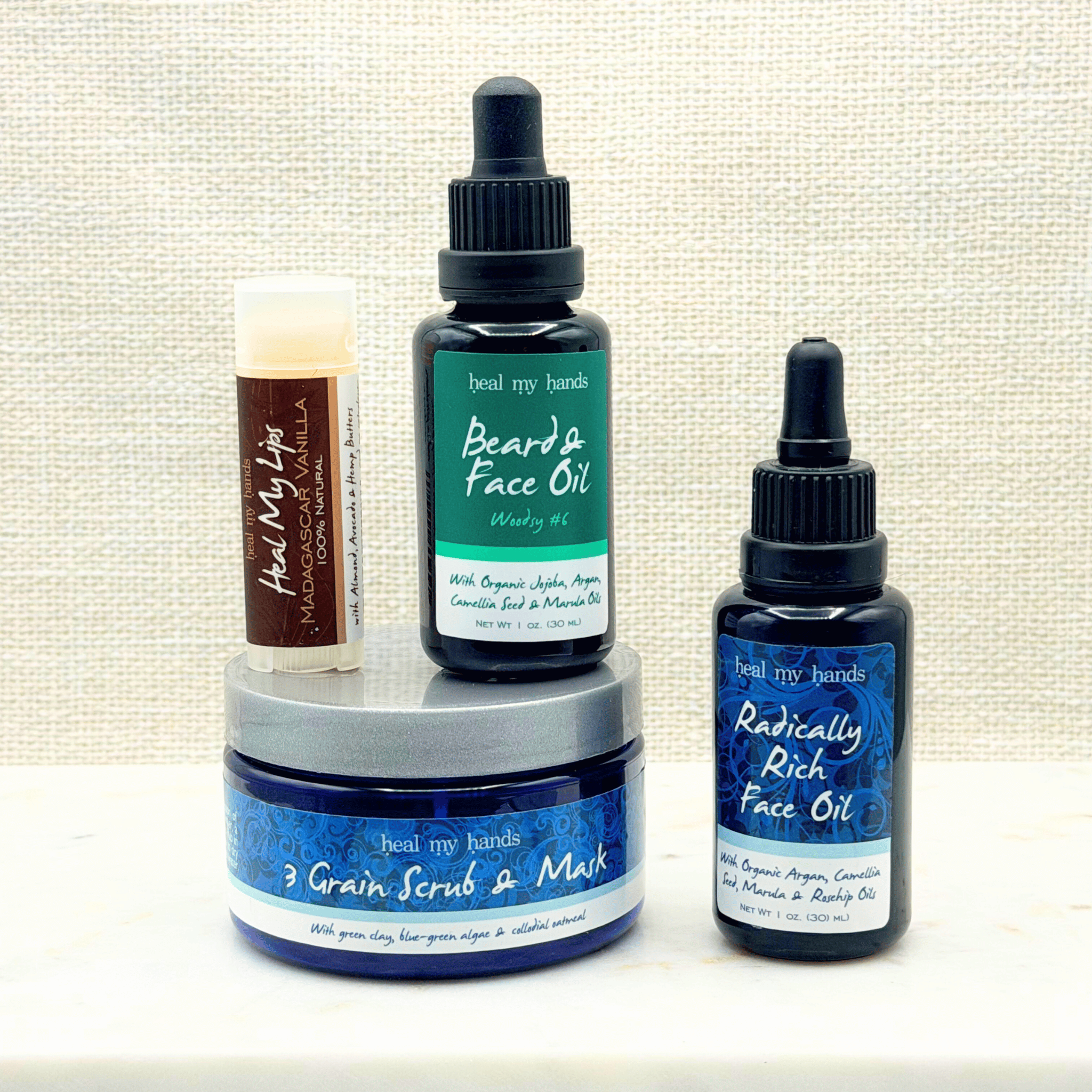 Collection of skincare products including a jar of 3 Grain Scrub & Mask, a bottle of Radically Rich Face Oil, a bottle of Beard & Face Oil, and a tube of Heal My Lips in Madagascar Vanilla on a marble surface with a textured beige background.