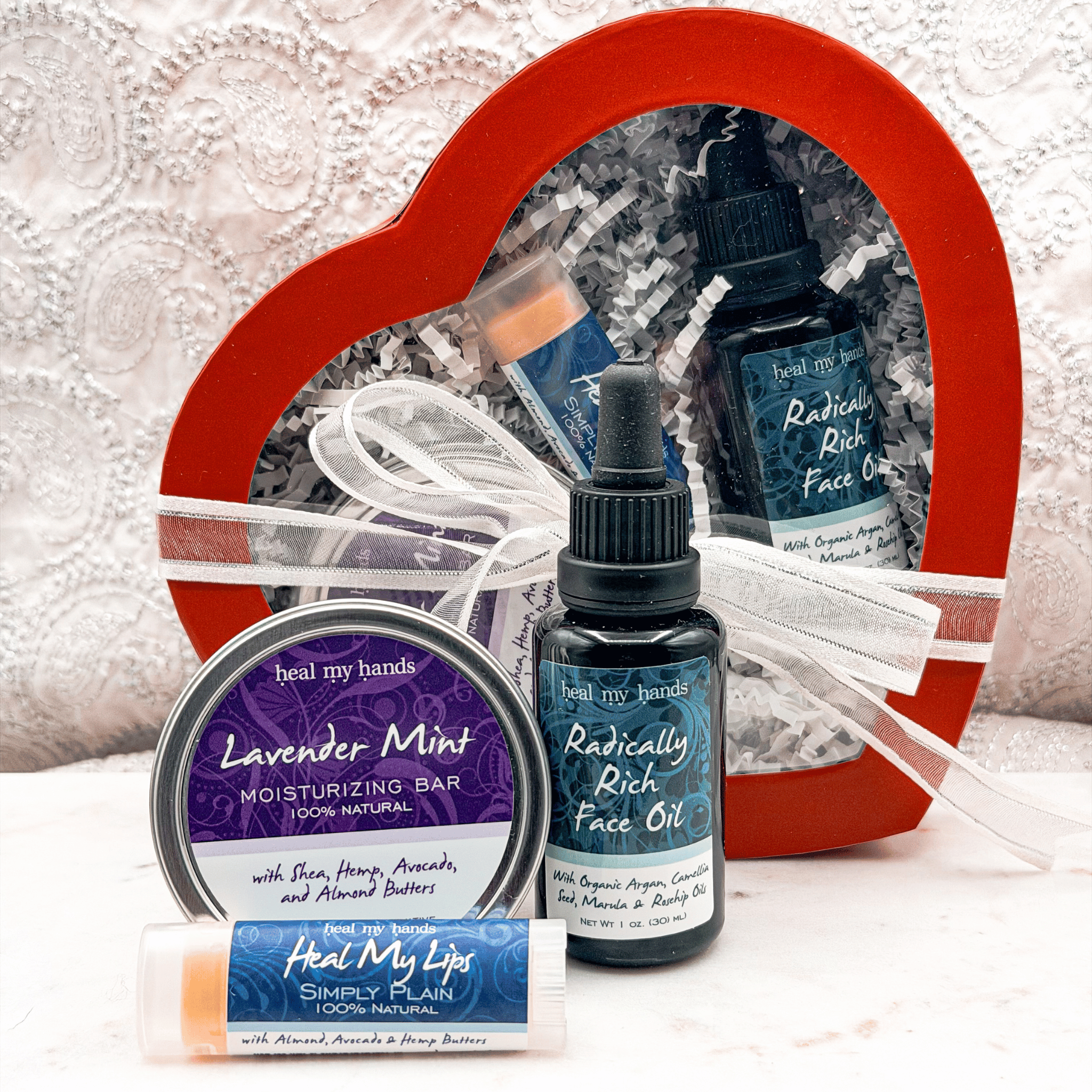 Heal My Hands skincare products including face oil, moisturizing bar, and lip balm with a heart-shaped gift box in the background.