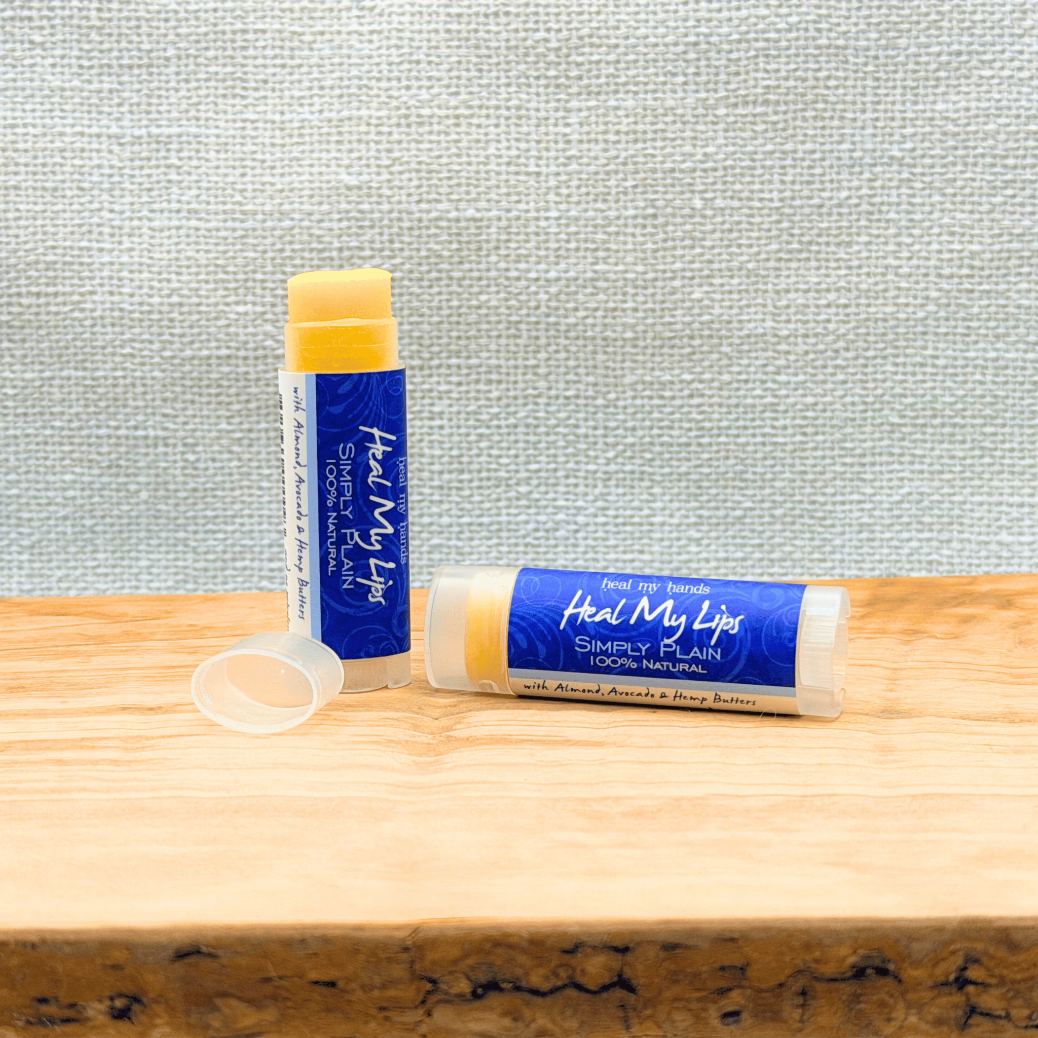 Two lip balm sticks with blue labels on a wooden surface against a textured gray wall.
