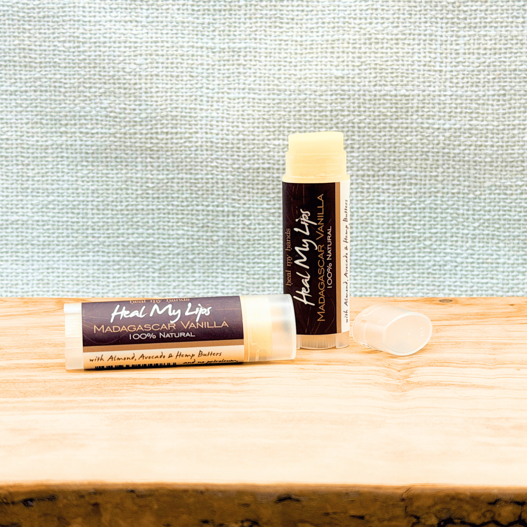 Two lip balms labeled 'Heal My Lips' on a wooden surface with a textured light blue background.