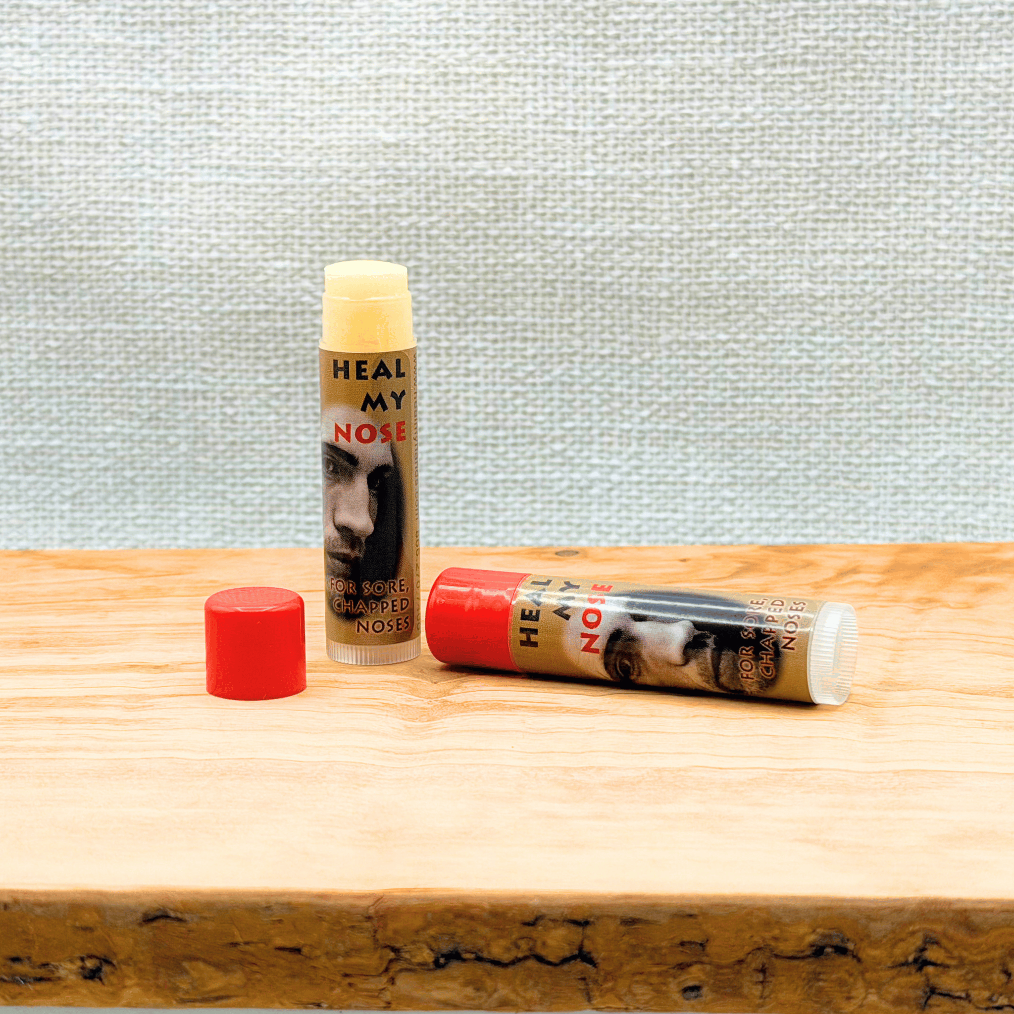 Two lip balm tubes labeled 'Heal My Nose' on a wooden surface with a textured light blue wall background.
