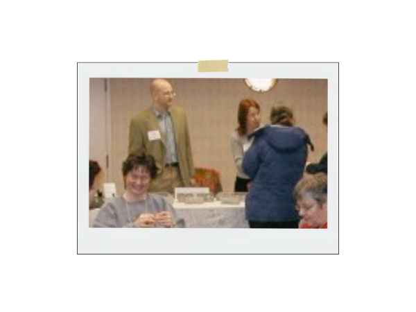 Group of people in a social setting, possibly a conference or event, with a focus on a woman sitting at a table.