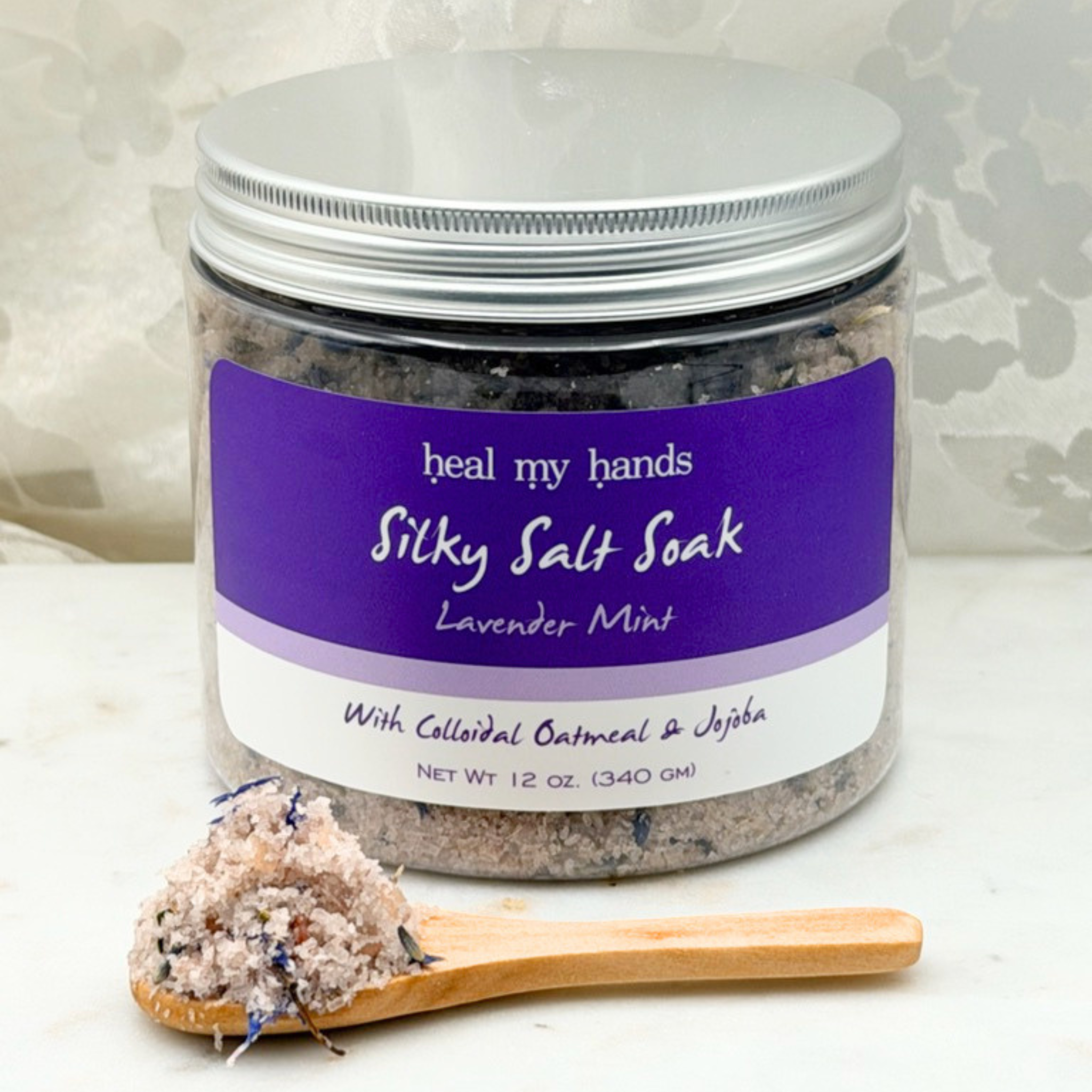 Jar of Lavender Mint Silky Salt Soak with small amount of bath salts on wooden spoon on marble surface.