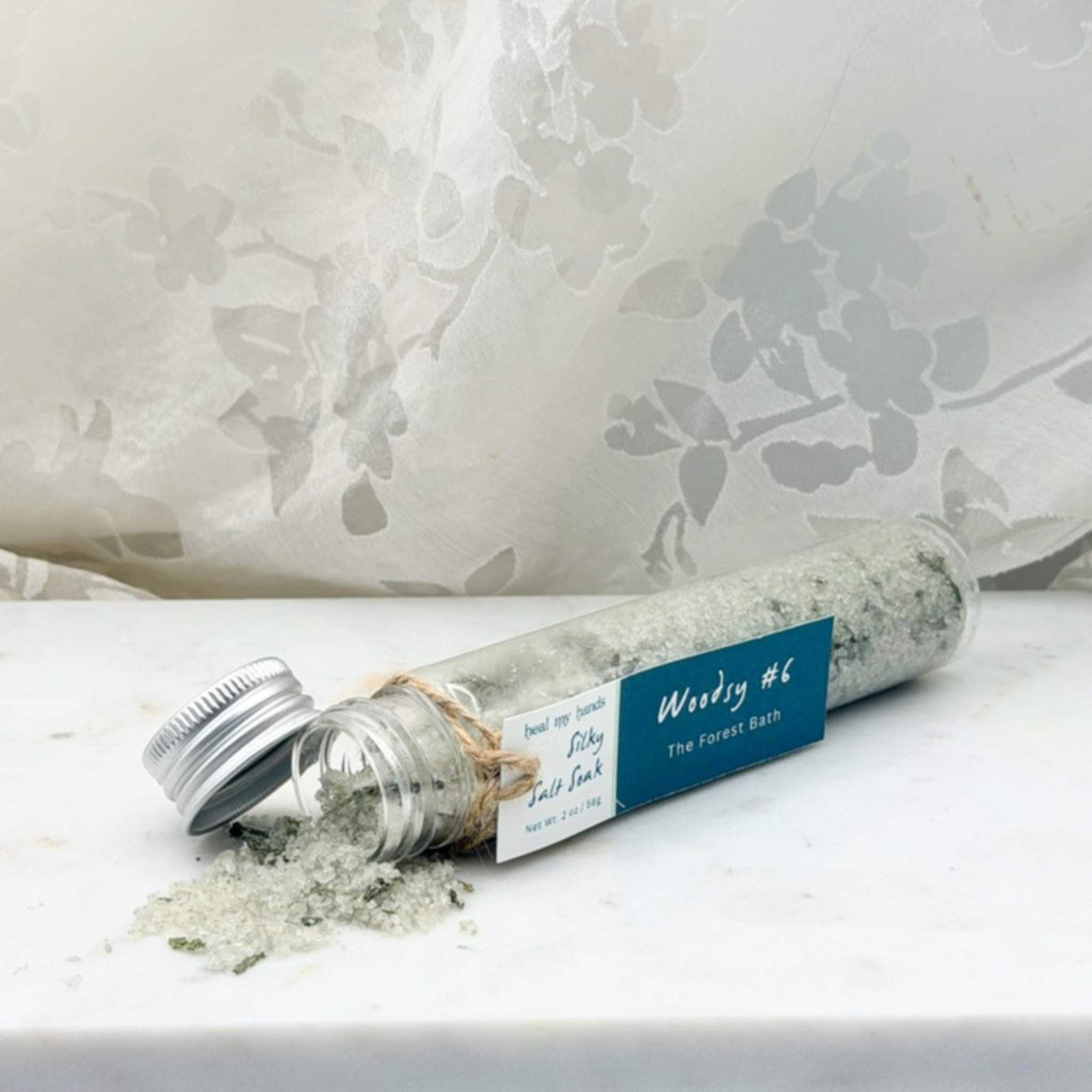 Woodsy #6 Silky Salt Soak spilling out of vial on marble surface.