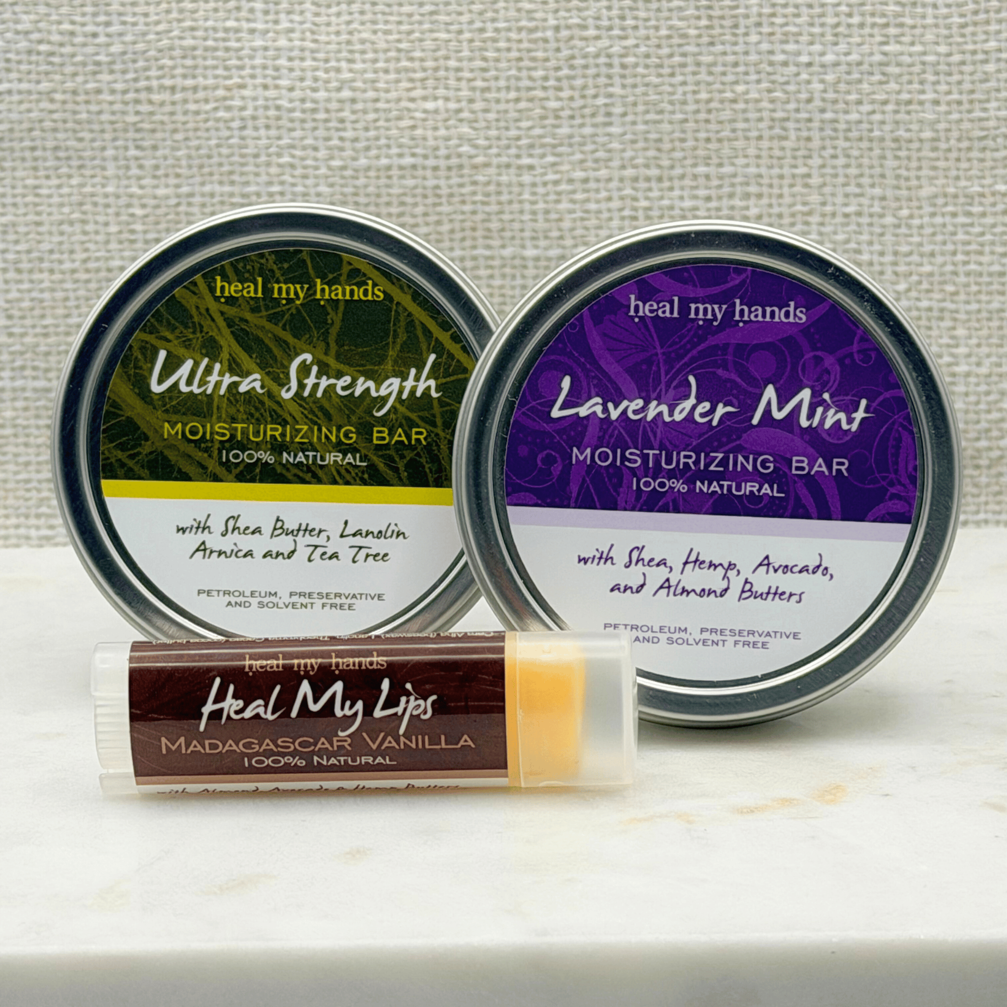 One tin of Ultra Strength Moisturizing Bar, one tin Lavender Mint Moisturizing Bar and one Heal My Lips in Madagascar Vanilla on a white marble surface with a white linen background. products from 'Heal My Hands' on a light background