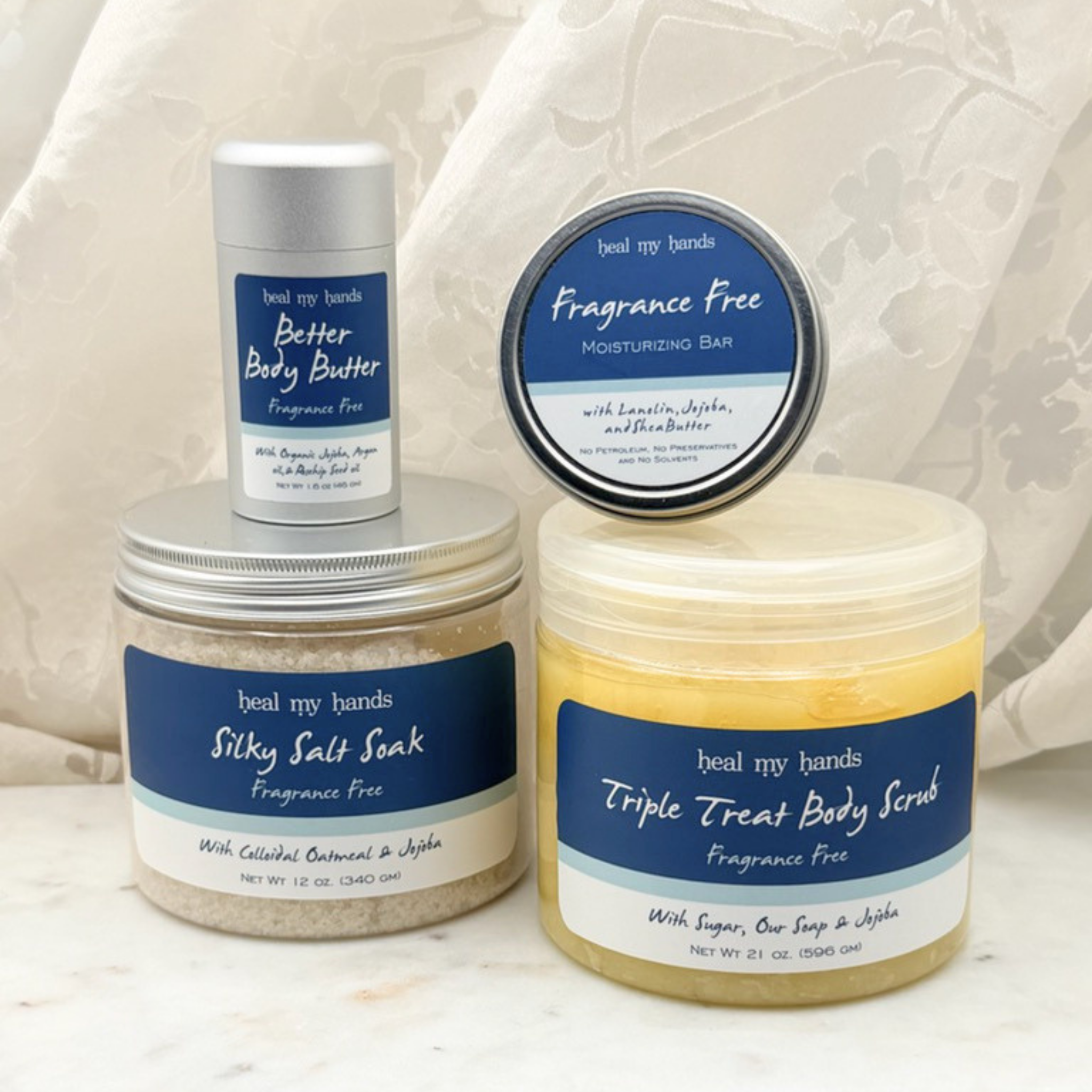 Collection of products, including a jar of Triple Treat Body Scrub, a jar of Silky Salt Soak, a tin of regular strength moisturizing bar, a tube of Better Body Butter on a white marble surface with a white curtain backdrop.