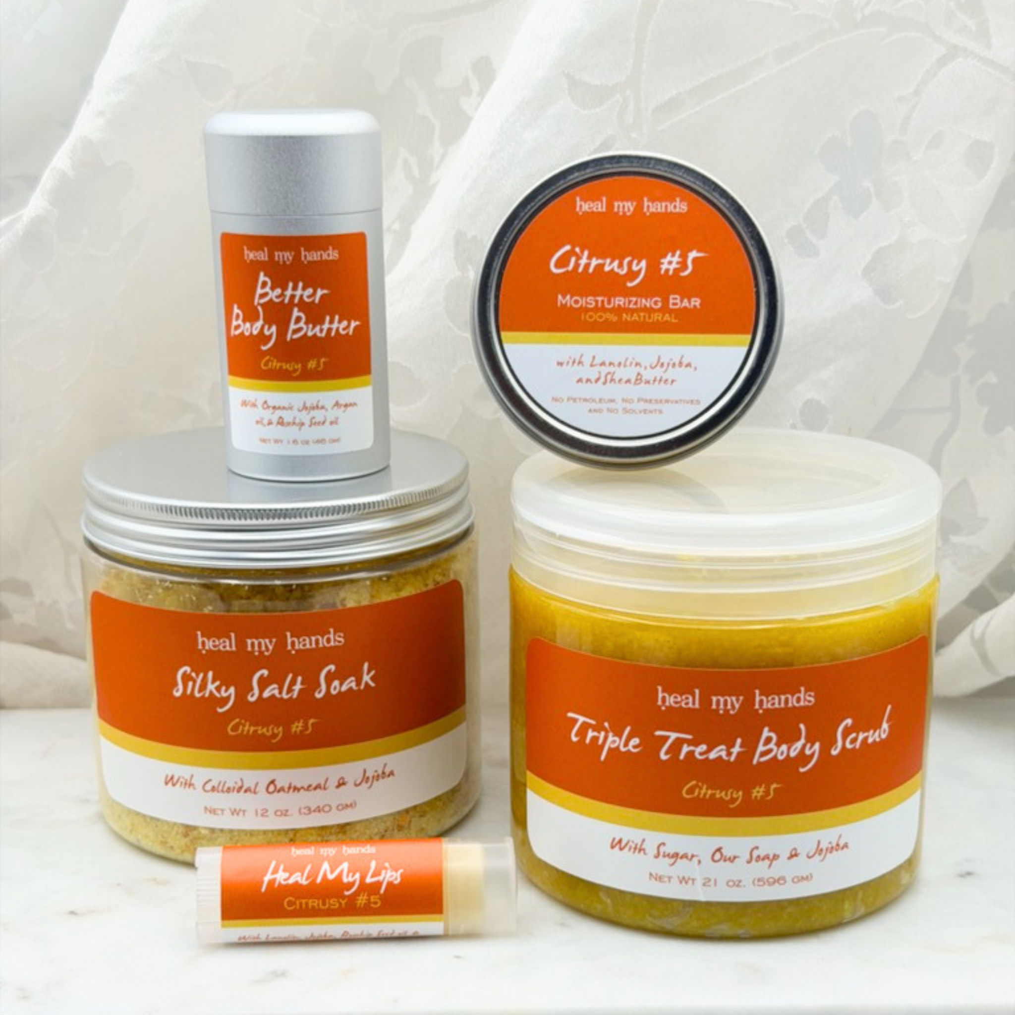 Collection of products, including a jar of Triple Treat Body Scrub, a jar of Silky Salt Soak, a tube of Heal My Lips, a tin of regular strength moisturizing bar, a tube of Better Body Butter on a white marble surface with a white curtain backdrop.