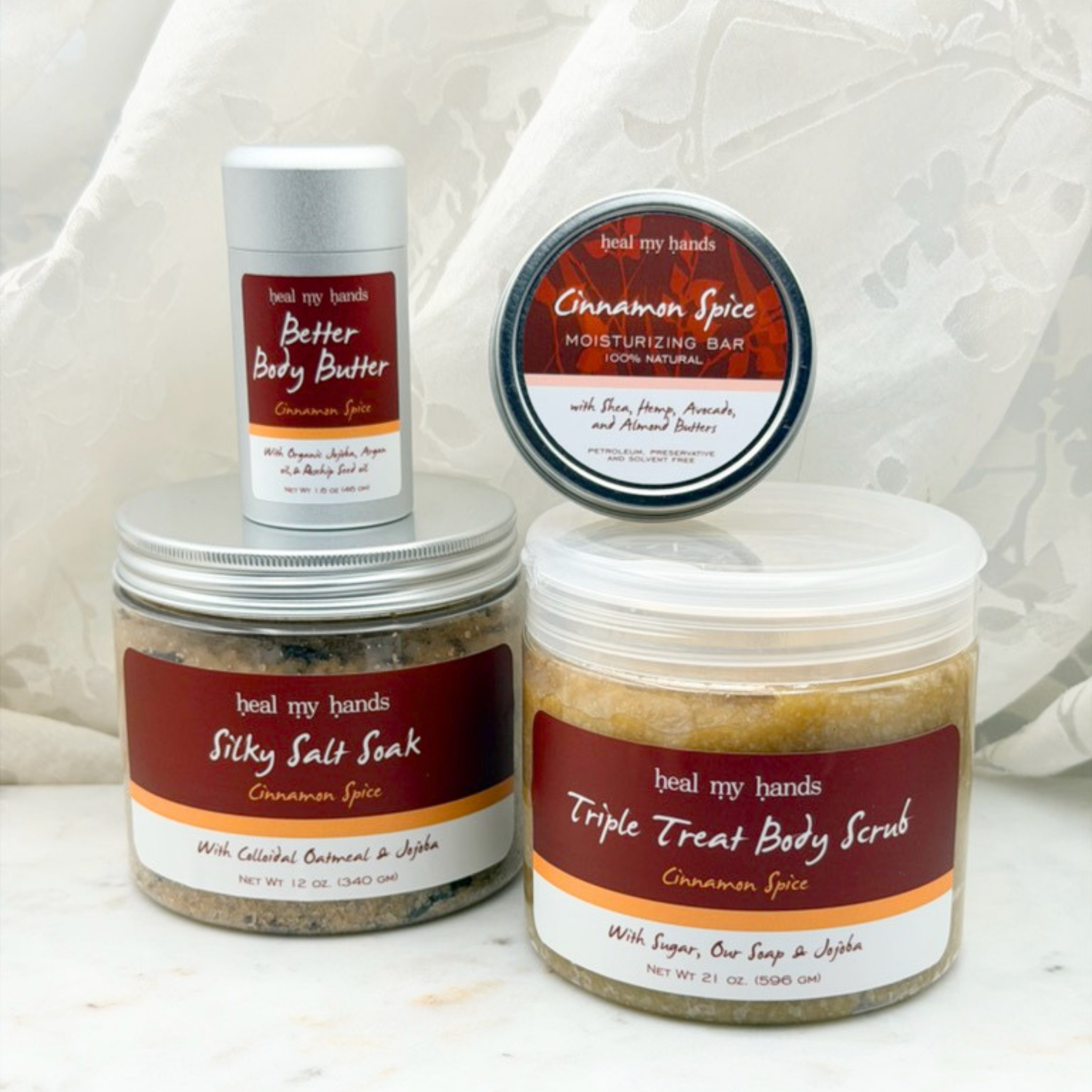 Collection of products, including a jar of Triple Treat Body Scrub, a jar of Silky Salt Soak, a tin of regular strength moisturizing bar, a tube of Better Body Butter on a white marble surface with a white curtain backdrop.