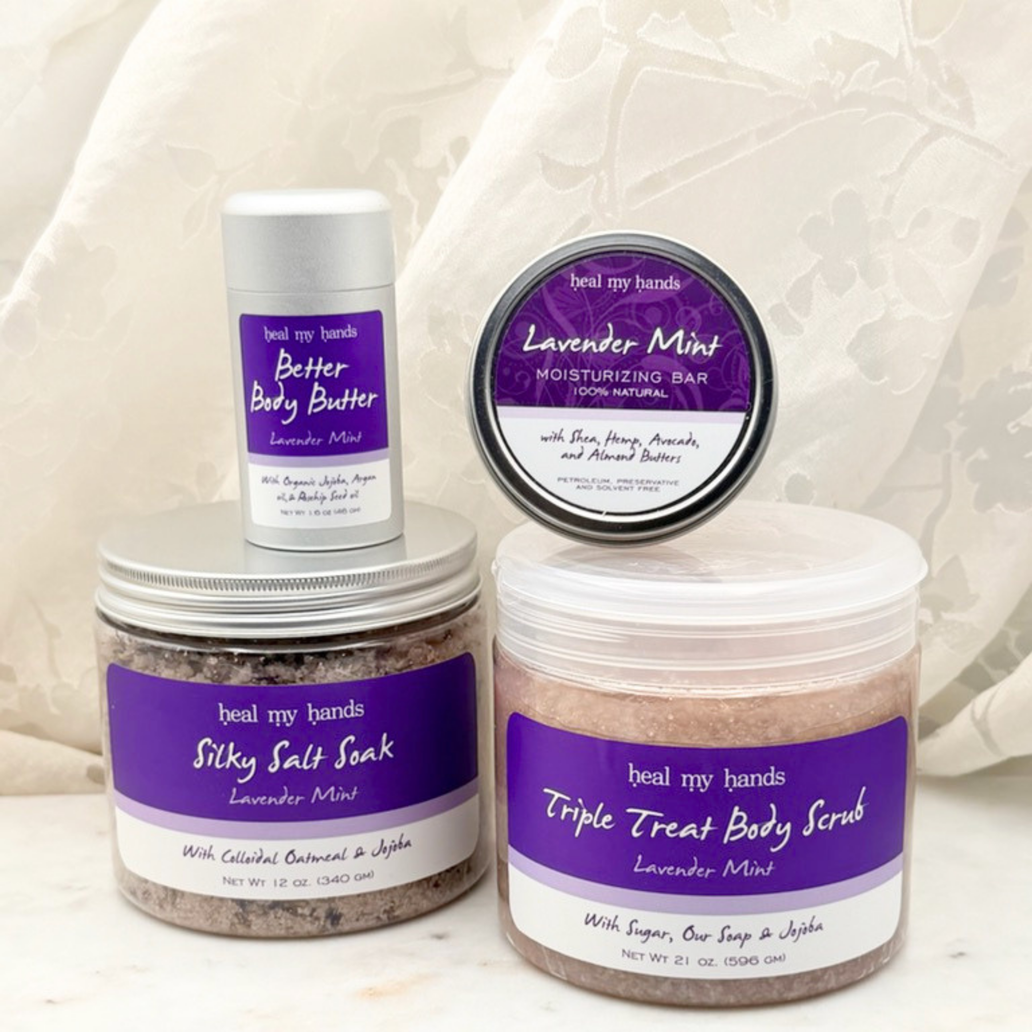 Collection of products, including a jar of Triple Treat Body Scrub, a jar of Silky Salt Soak, a tin of regular strength moisturizing bar, a tube of Better Body Butter on a white marble surface with a white curtain backdrop.