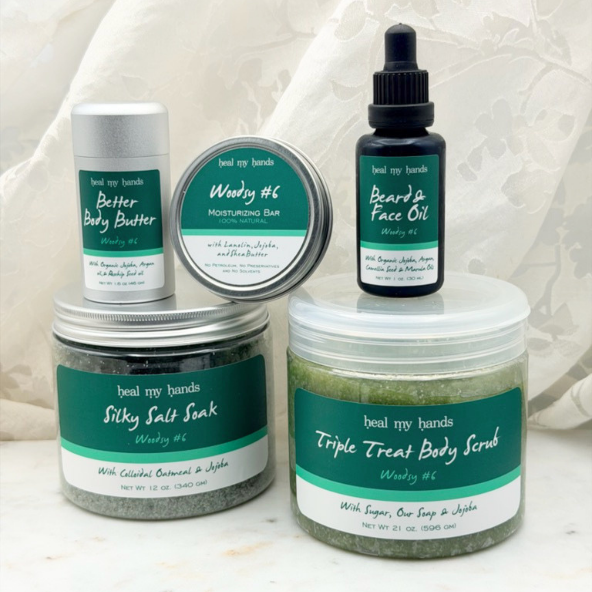 Collection of products, including a jar of Triple Treat Body Scrub, a jar of Silky Salt Soak, a bottle of Beard & Face Oil, a tin of regular strength moisturizing bar, a tube of Better Body Butter on a white marble surface with a white curtain backdrop.