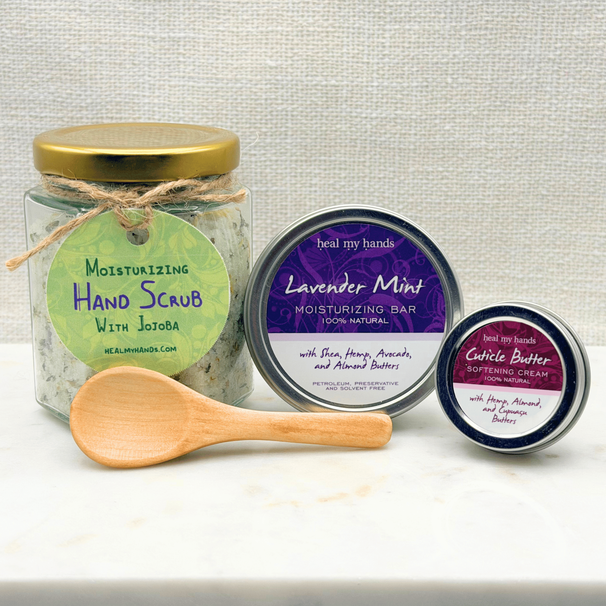 Set of natural skincare products including a jar of hand scrub, a moisturizing bar in Lavender Mint scent and a tin of cuticle butter on a marble surface with a cream textured background.