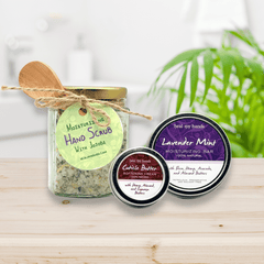 Three natural skincare products on a wooden surface with a plant in the background. Moisturizing Hand Scrub, HealMy Hands Moisturizing Bar in Lavender Mint and Cuticle Butter.