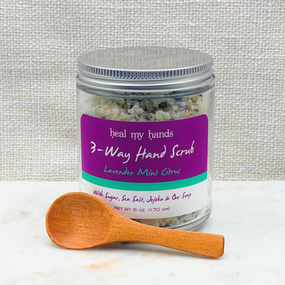 A jar of 'heal my hands' 3-Way Hand Scrub with a purple label, shown with a small wooden spoon on a white marble surface against a textured beige fabric background.
