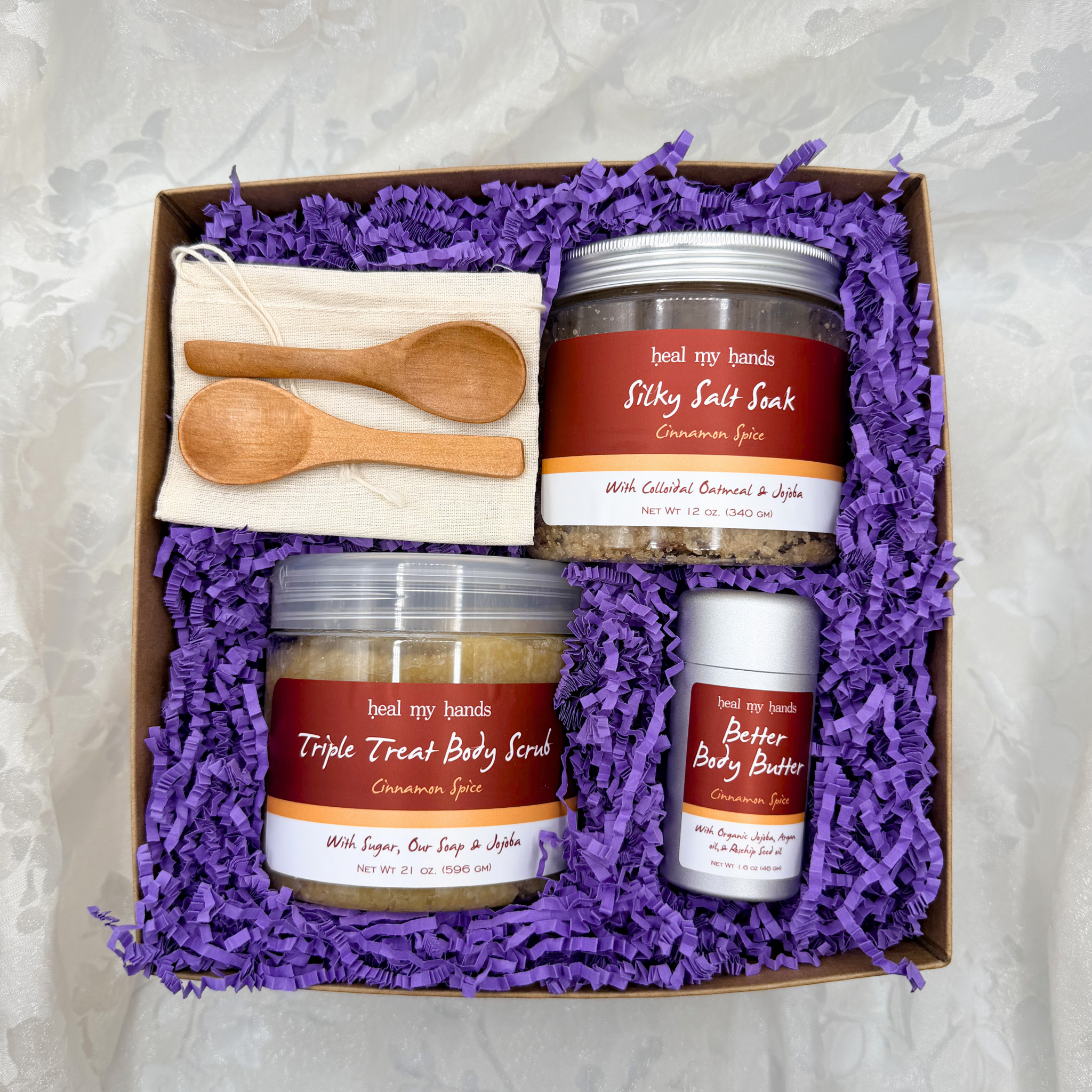 Box filled with purple crinkle paper and a jar of Silky Salt Soak, Triple Treat Body Scrub, and Better Body Butter in Cinnamon Spice with two small wooden spoons and a muslin pouch.