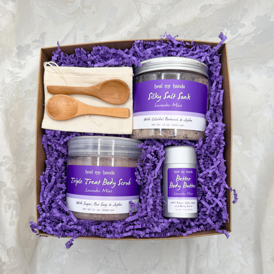 Box filled with purple crinkle paper and a jar of Silky Salt Soak, Triple Treat Body Scrub, and Better Body Butter in Lavender Mint with two small wooden spoons and a muslin pouch.