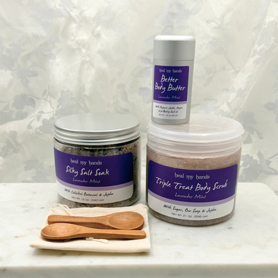 A jar of Silky Salt Soak, Triple Treat Body Scrub, and Better Body Butter in Lavender Mint with two small wooden spoons and a muslin pouch on a marble surface.