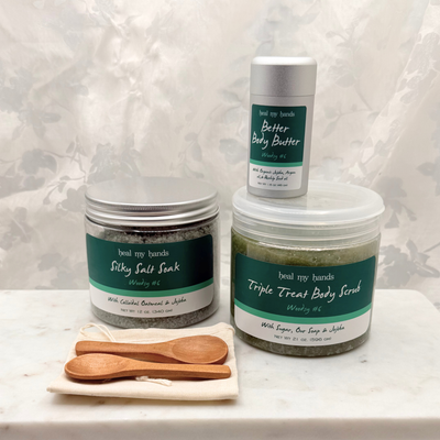 A jar of Silky Salt Soak, Triple Treat Body Scrub, and Better Body Butter in Woodsy #6 with two small wooden spoons and a muslin pouch on a marble surface.