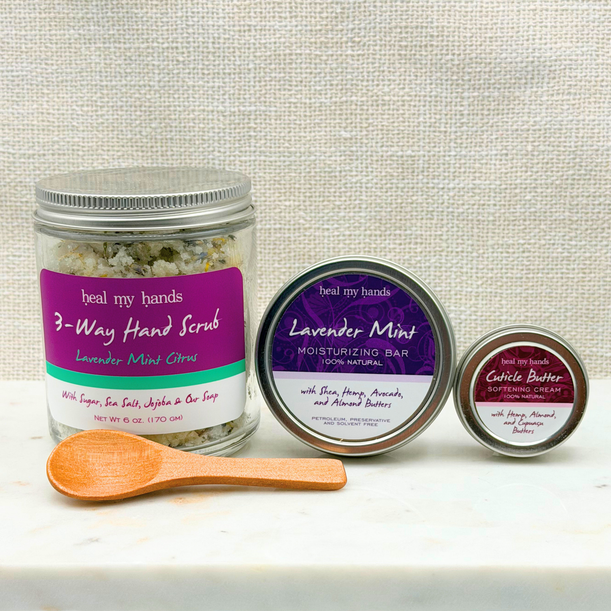 Three containers of 'heal my hands' hand care products and a wooden spoon on a white marble surface against a textured beige fabric background. The products include a large jar of 3-Way Hand Scrub, a round tin of Lavender Mint Moisturizing Bar, and a small tin of Cuticle Butter.