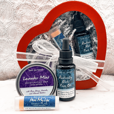 Heal My Hands skincare products including face oil, moisturizing bar, and lip balm with a heart-shaped gift box in the background.