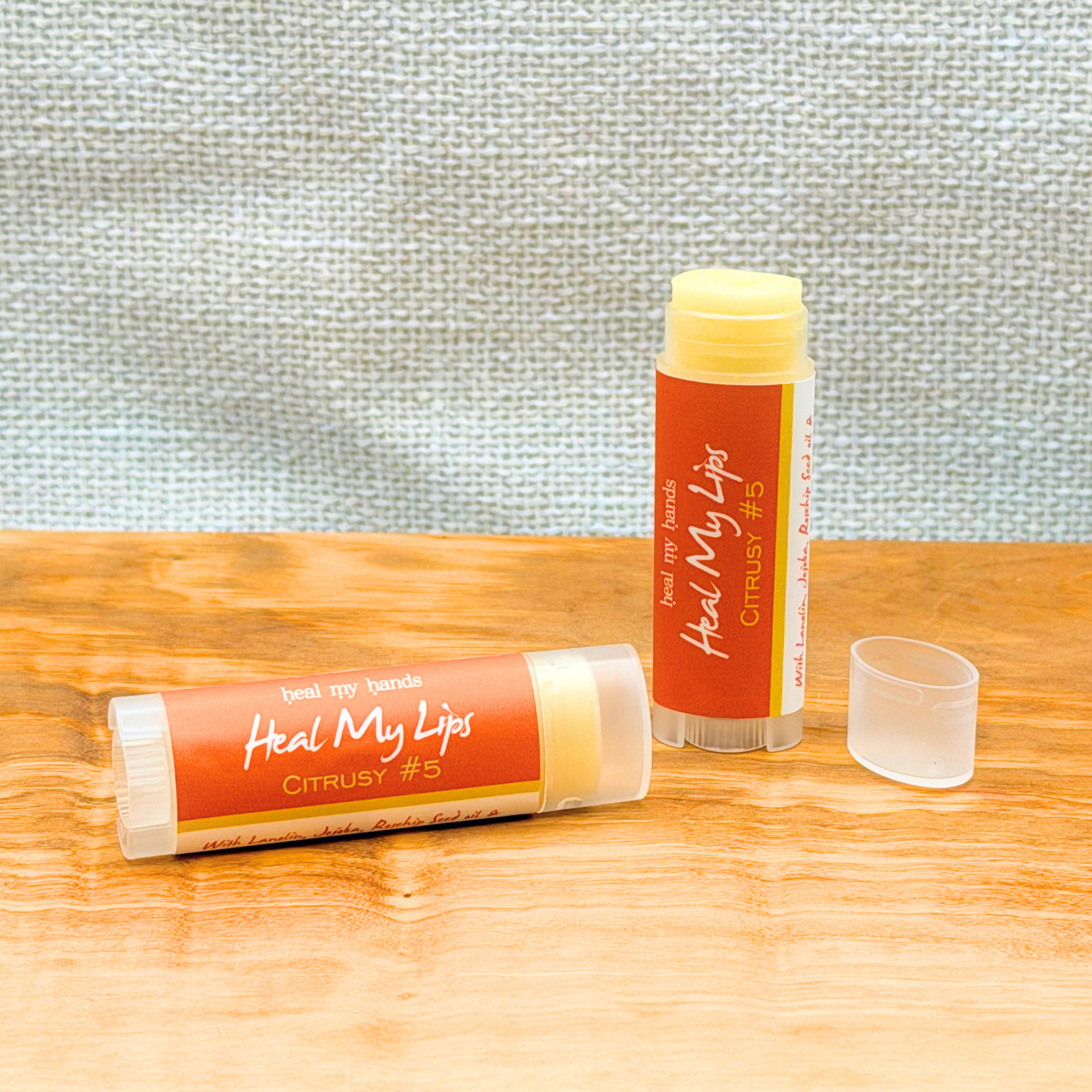 Two Citrusy #5 lip balm tubes with orange labels on a wooden surface against a textured gray wall.