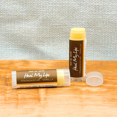 Two lip balm tubes with brown labels on a wooden surface against a textured gray wall.