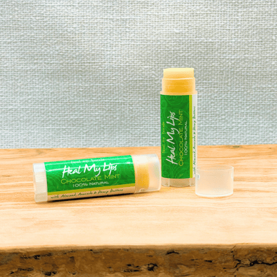 Two lip balm tubes with green labels on a wooden surface against a textured gray wall.