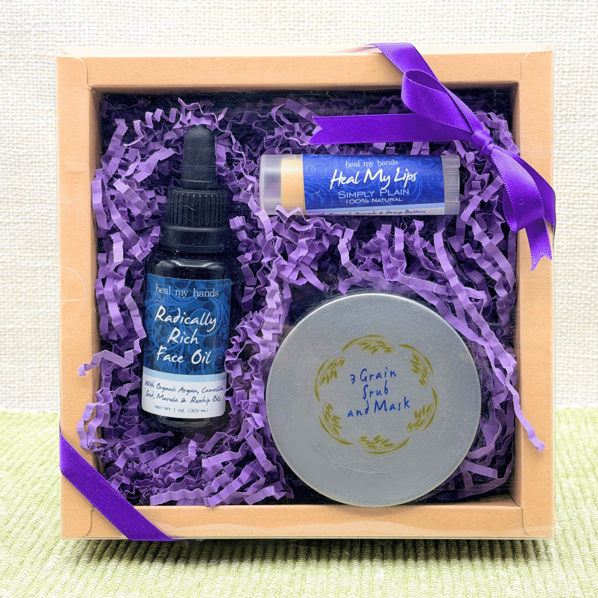 Skincare product set with face oil, lip balm, and scrub in a box with purple ribbon and filler