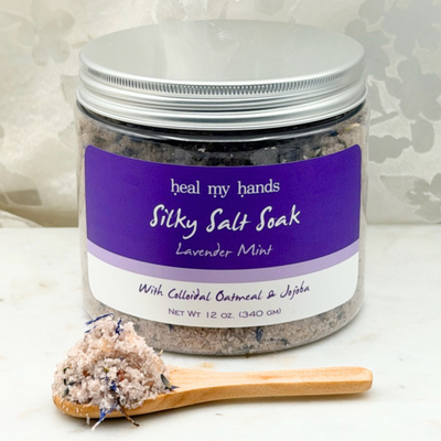 Jar of Lavender Mint Silky Salt Soak with small amount of bath salts on wooden spoon on marble surface.