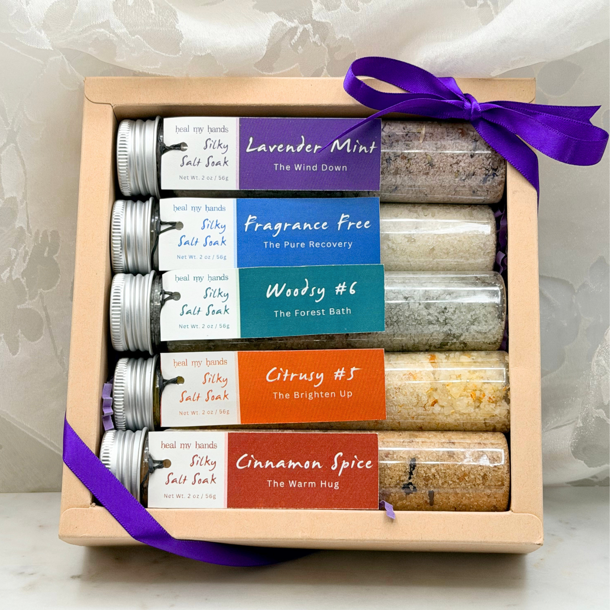 Box of 5 vials of Silky Salt Soak tied with a purple ribbon on a white marble surface.