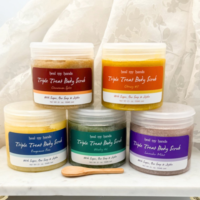 5 jars of Triple Treat Body Scrub in Cinnamon Spice, Citrusy #5, Fragrance Free, Woodsy #6 and Lavender Mint with a small wooden spoon on marble surface.