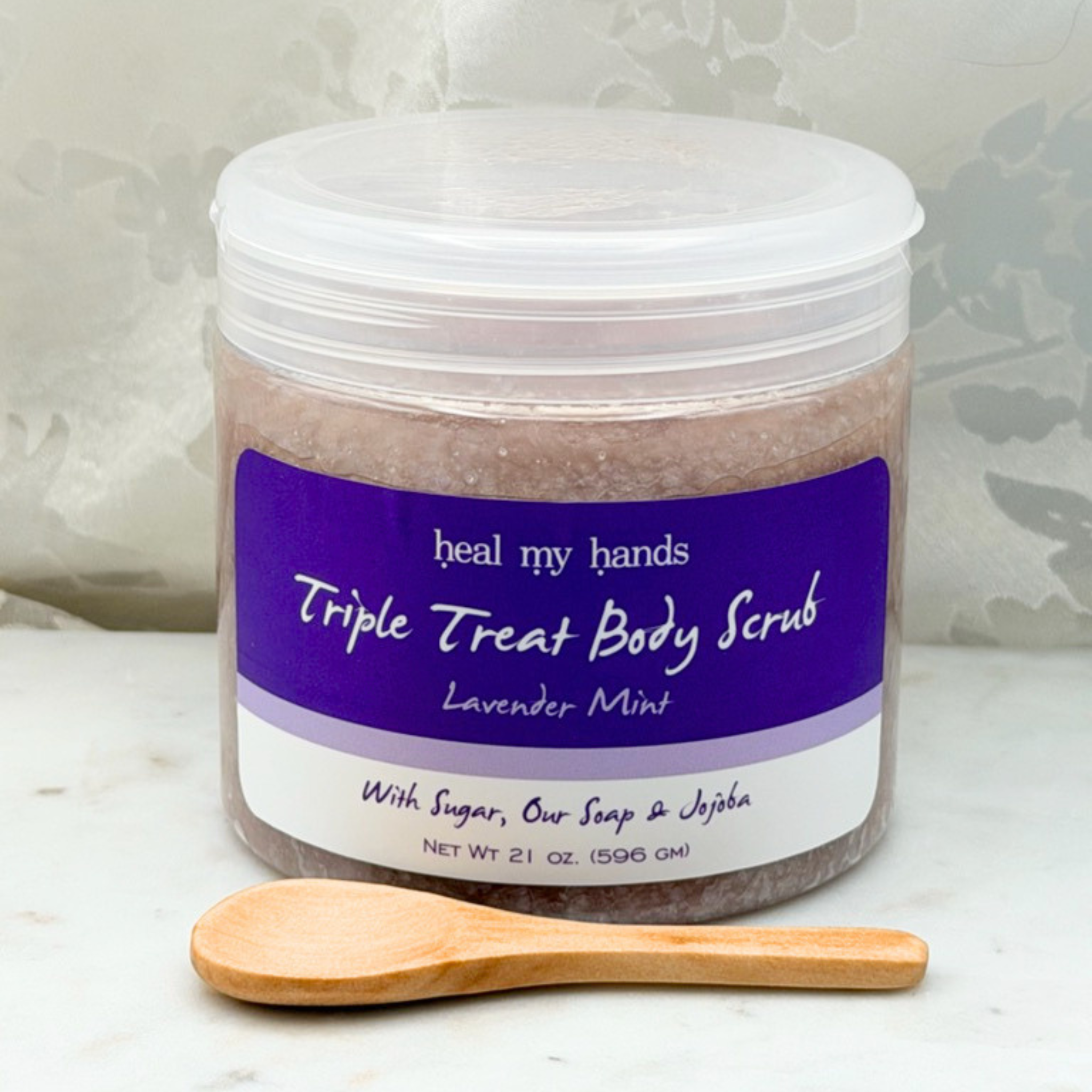Lavender Mint Triple Treat Body Scrub jar and small wooden spoon on marble surface.